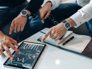 Business professionals analysing financial charts on a tablet, discussing hidden marketing costs affecting investment decisions.