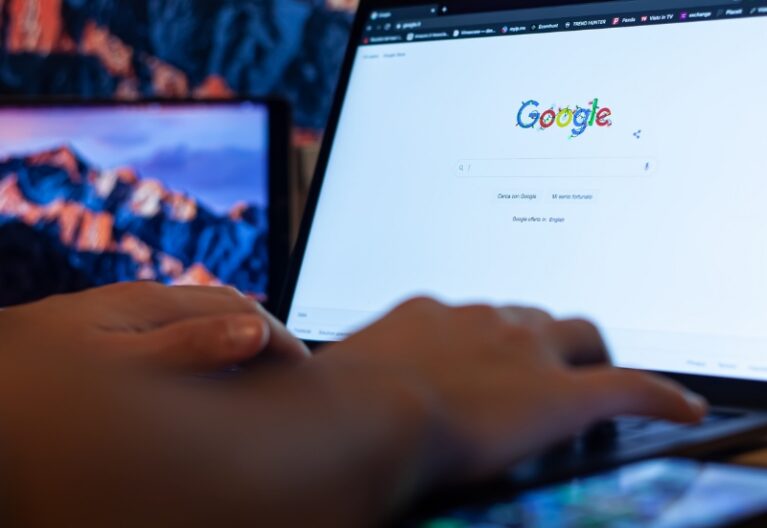 ai-focused view of hands typing on laptop showing Google homepage, with smartphone in foreground and monitors behind.