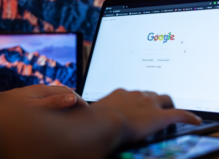 ai-focused view of hands typing on laptop showing Google homepage, with smartphone in foreground and monitors behind.