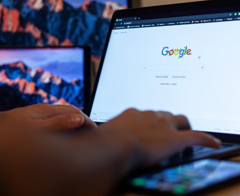 ai-focused view of hands typing on laptop showing Google homepage, with smartphone in foreground and monitors behind.