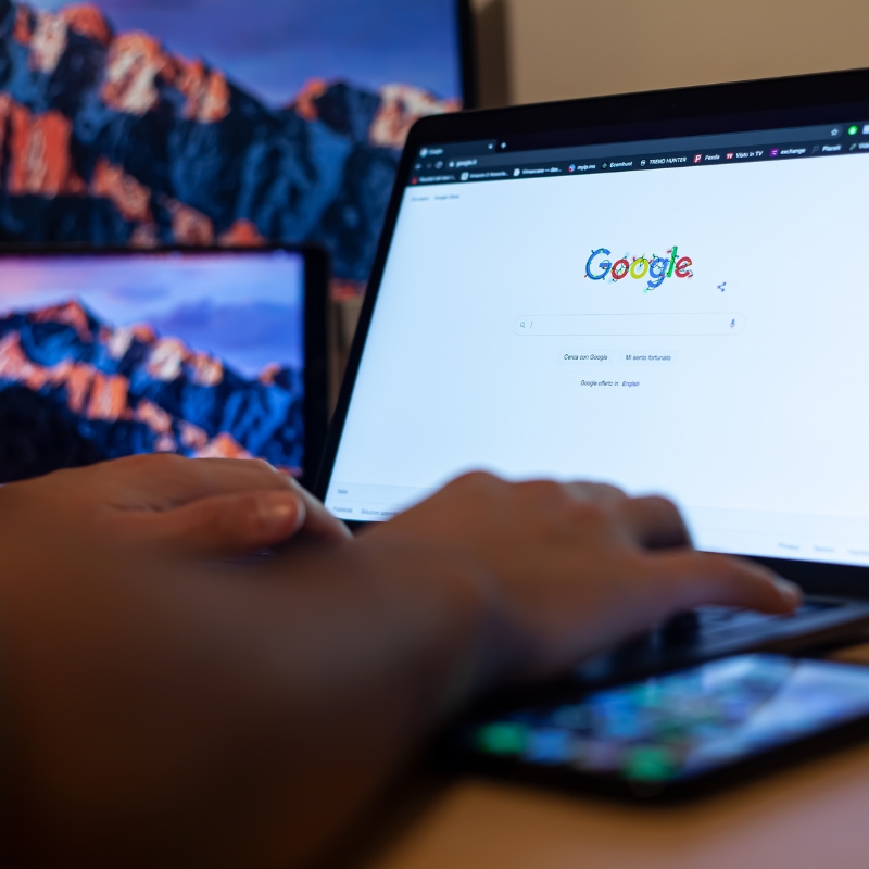 ai-focused view of hands typing on laptop showing Google homepage, with smartphone in foreground and monitors behind.