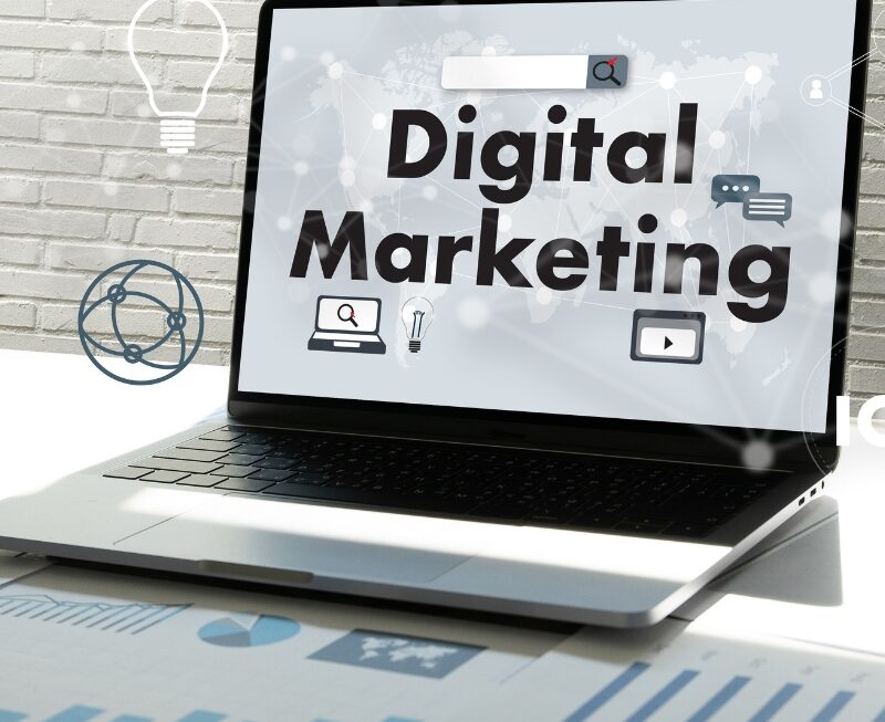 Laptop screen displaying bold "Digital Marketing" text surrounded by online icons on a office desk setup.